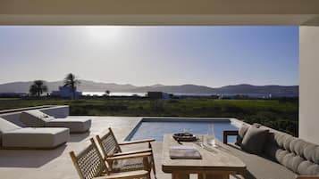 A poolside area with a table, chairs, and a sofa, overlooking a body of water and distant mountains.