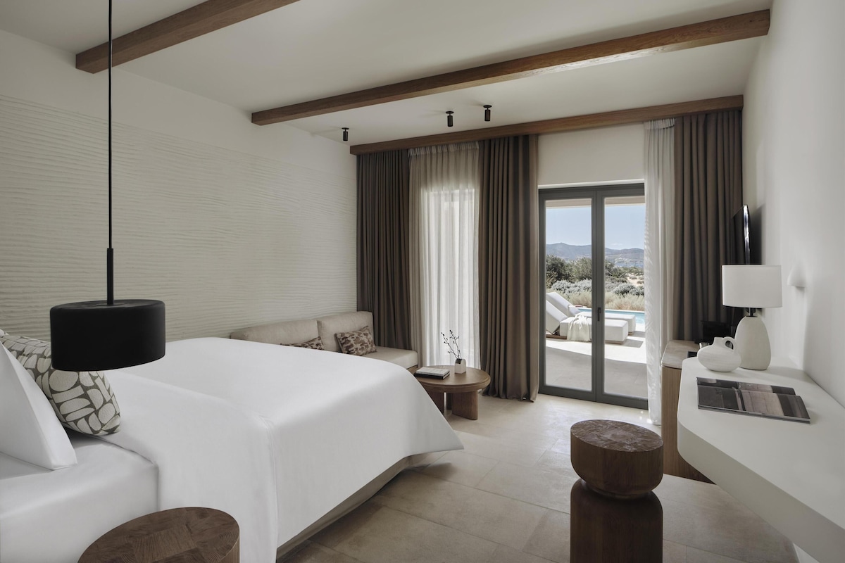 Habitación de hotel con cama, sofá, una mesita, televisor y vistas a una piscina y al paisaje.