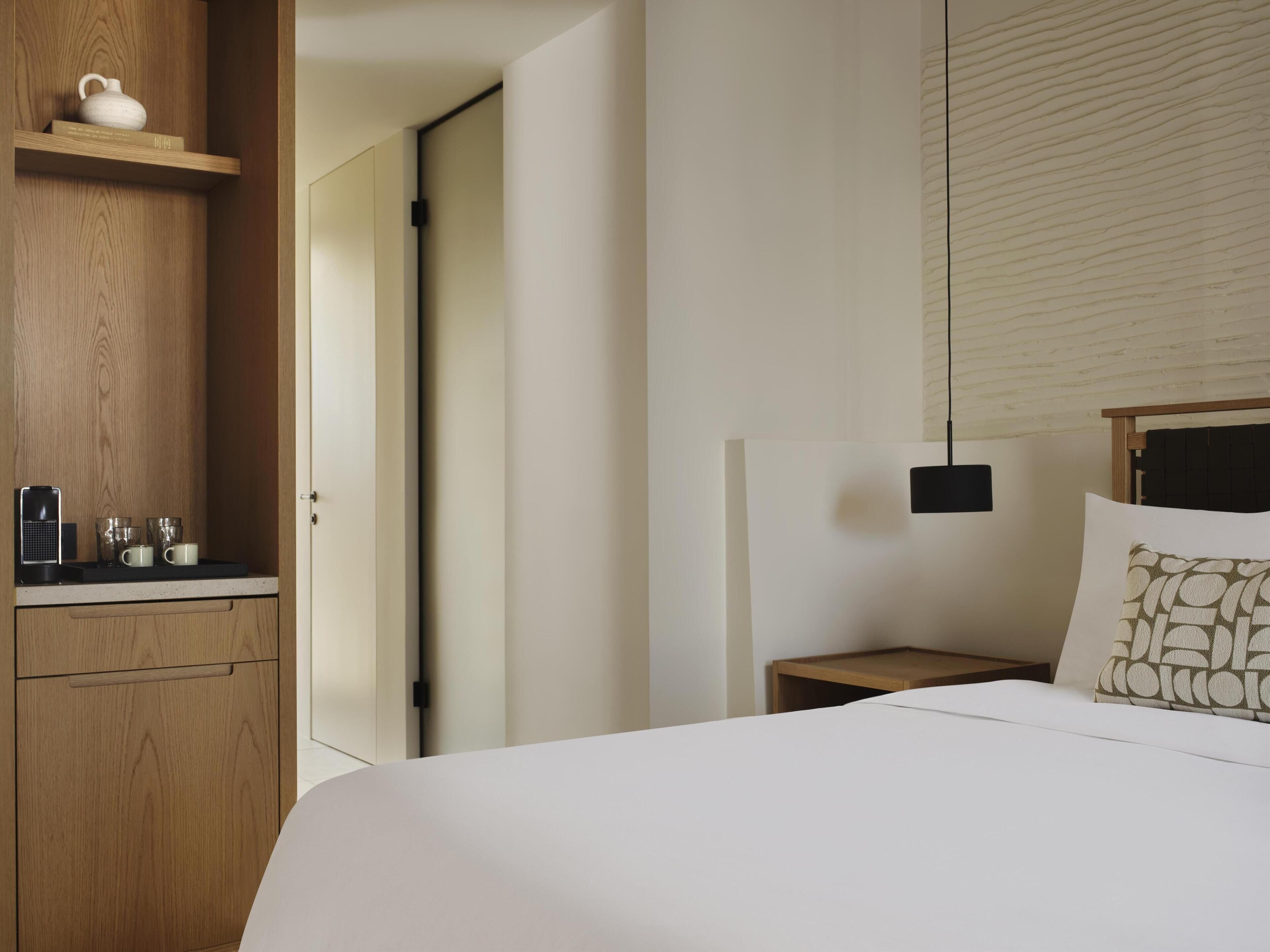 A hotel room with a wooden wardrobe, a bedside table with a lamp, a bed with a patterned pillow, and a window with blinds.