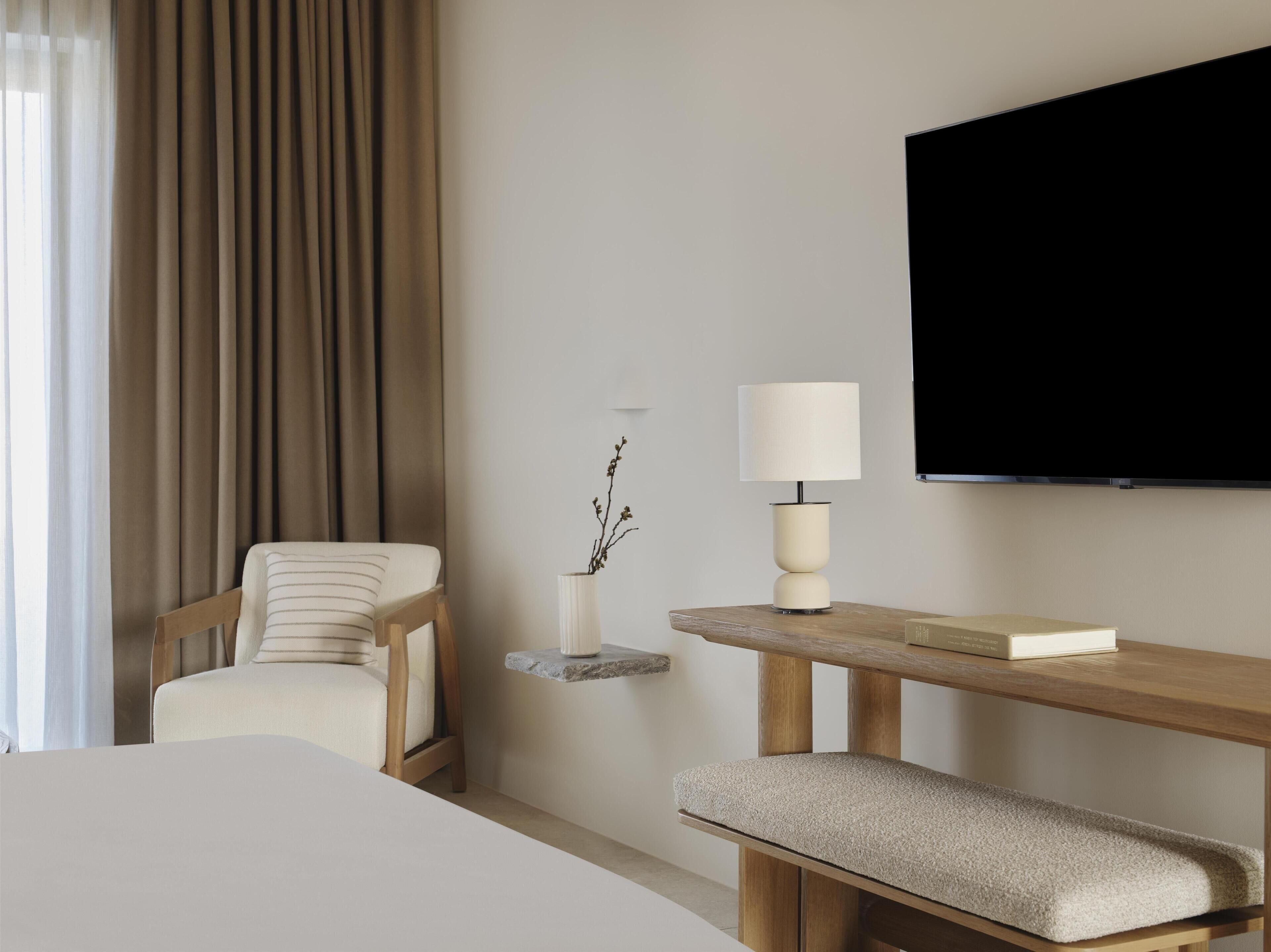 A hotel room with a bed, a chair, a bedside table with a lamp, a small shelf with a plant, and a flat-screen TV mounted on the wall.