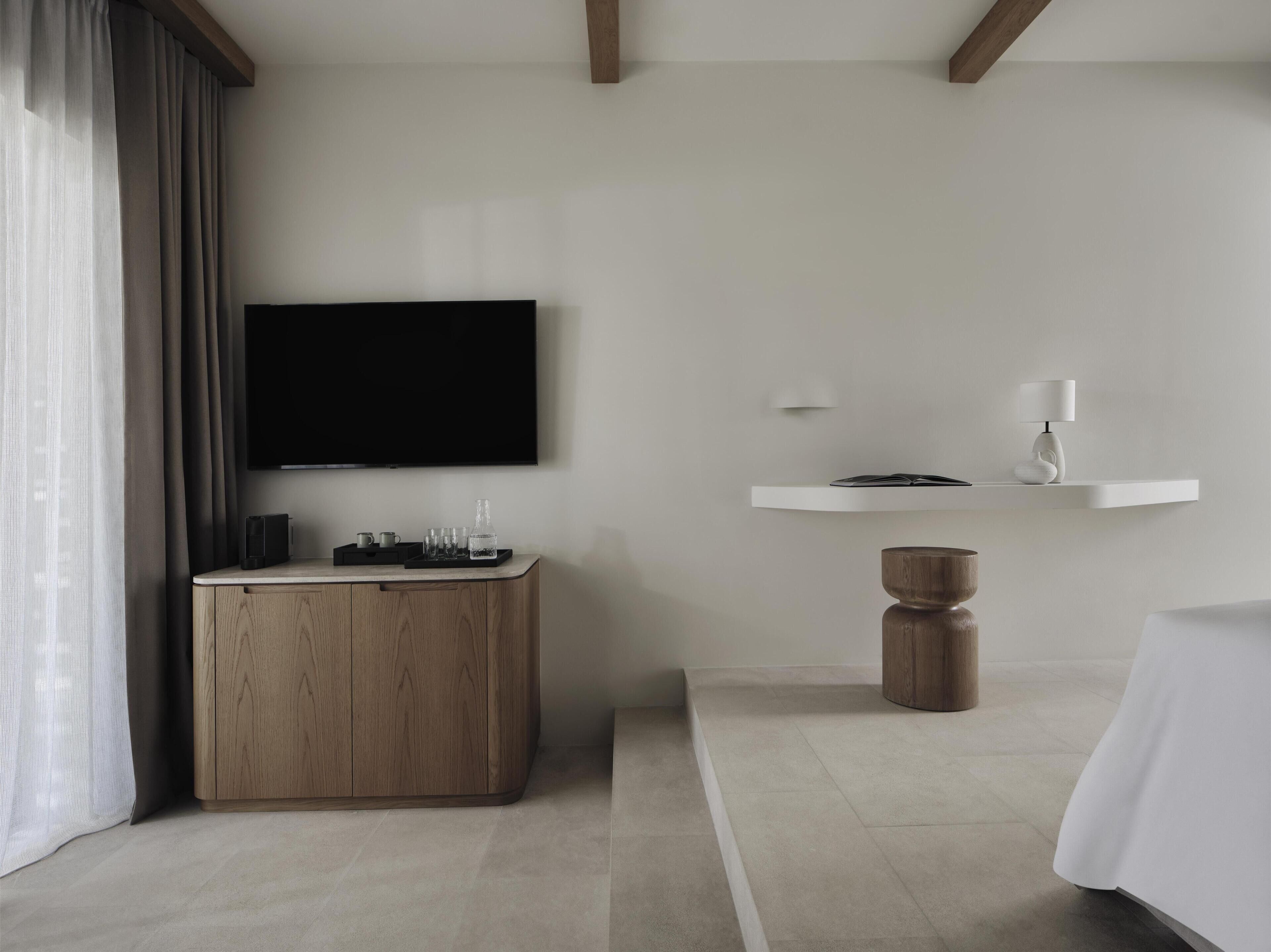 A modern hotel room with a flat-screen TV mounted on the wall, a wooden side table with a tray of glasses, a white bedside table with a lamp, and a bed with white bedding.