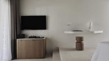 A modern hotel room with a flat-screen TV mounted on the wall, a wooden side table with a tray of glasses, a white bedside table with a lamp, and a bed with white bedding.