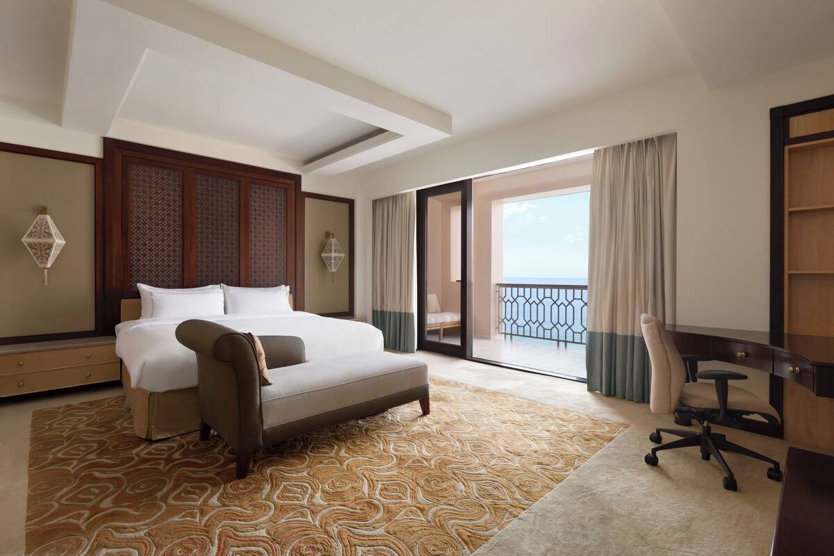 Une chambre d’hôtel avec un grand lit, un bureau, une chaise et une vue sur la mer depuis un balcon.