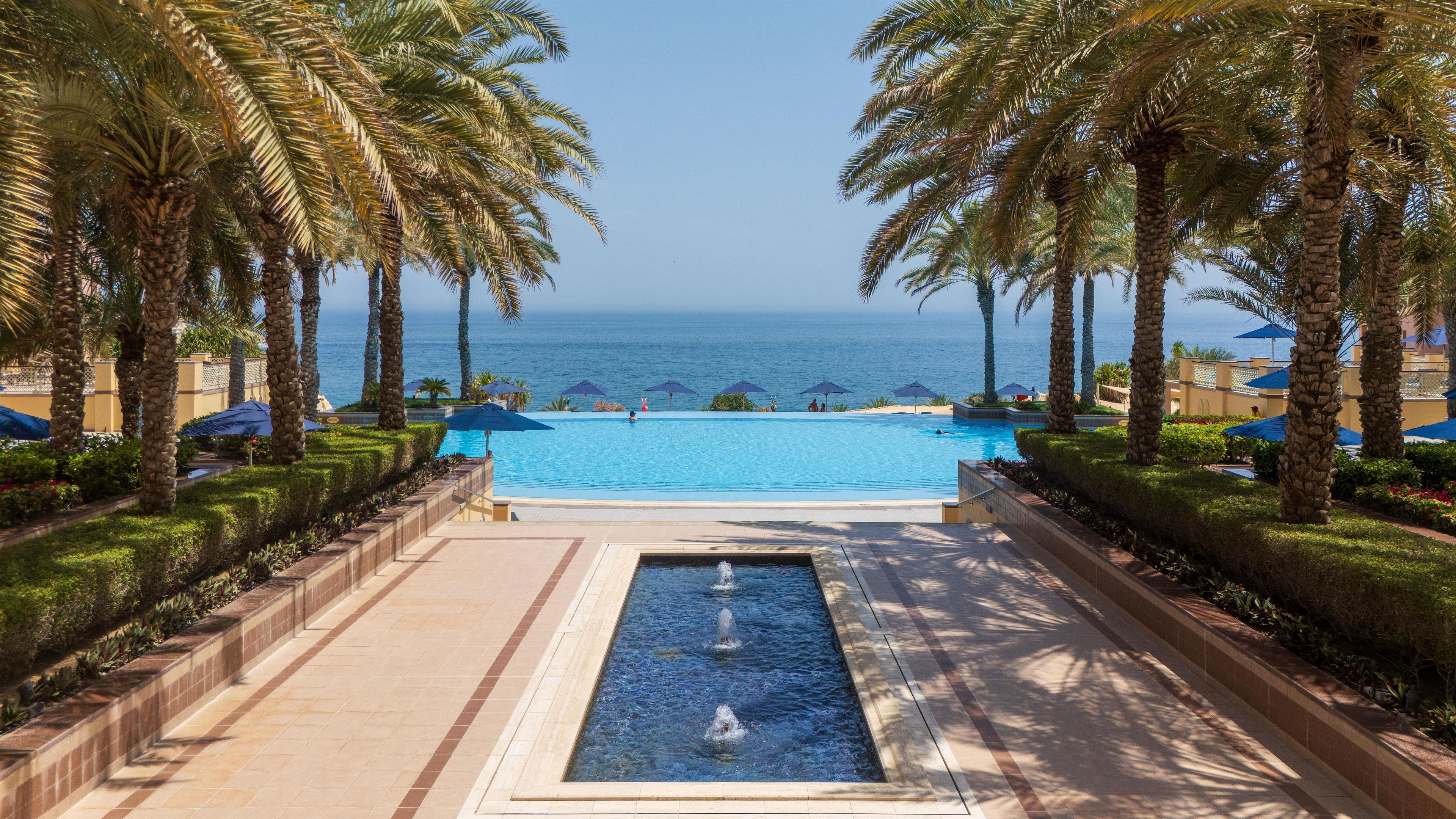 A pool with a view of the sea, surrounded by palm trees and umbrellas.