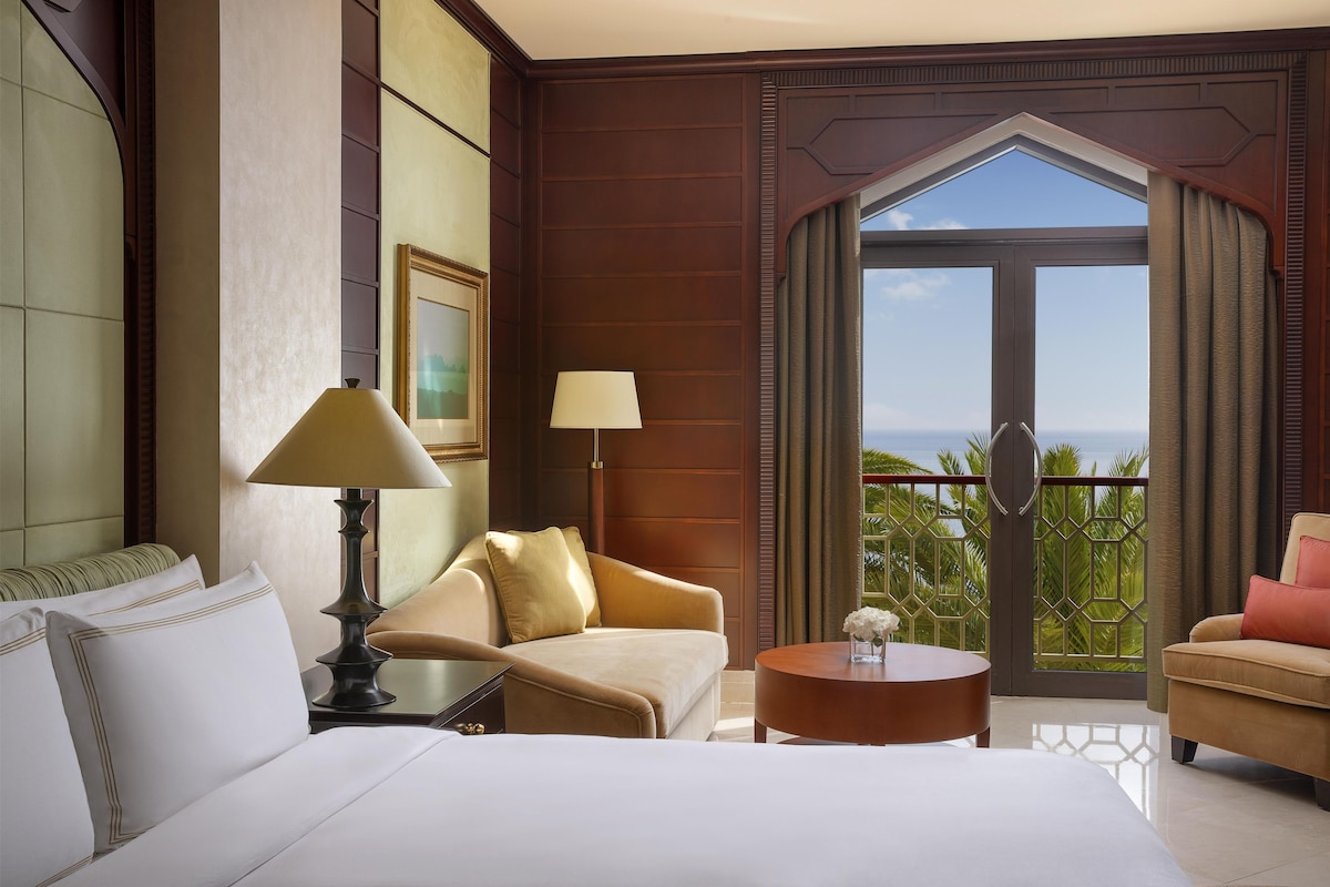 A hotel room with a bed, two chairs, a small table, a lamp, and a view of the ocean through a large door.