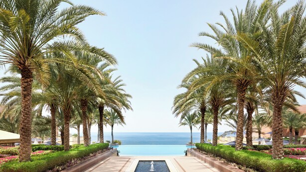 A pathway leading to a pool with palm trees on either side and a view of the ocean. - Al Husn Hotel Muscat - managed by Hilton (Muscat)