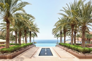 A pathway leading to a pool with palm trees on either side and a view of the ocean. - Al Husn Hotel Muscat (Muscat)
