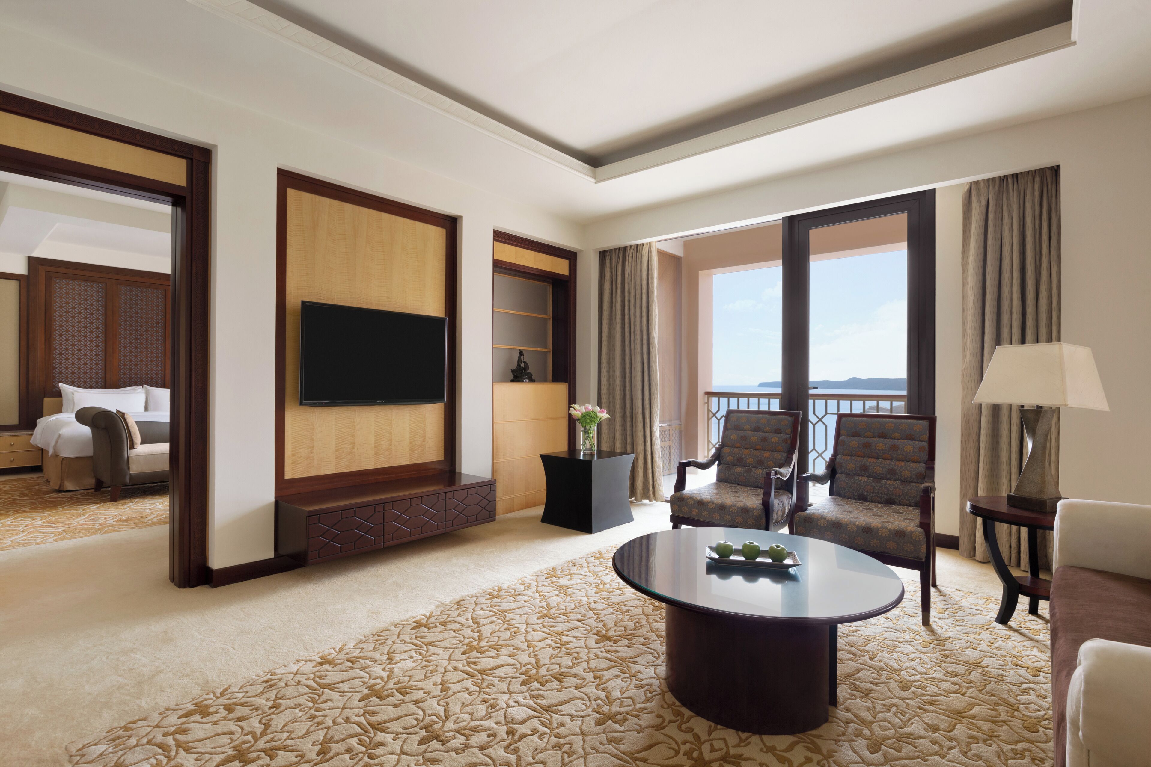 Een hotelkamer met een grote flatscreen televisie, een ronde salontafel, twee fauteuils, een bank, een decoratieve vaas en uitzicht op het omliggende landschap.