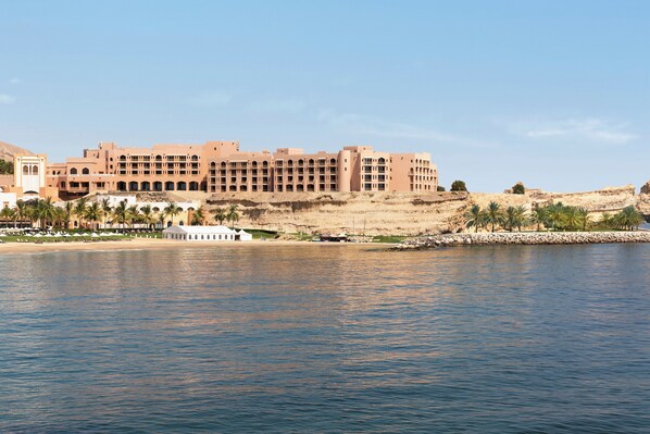 A large, multi-story building with a symmetrical facade, situated by a calm body of water. - Al Husn Hotel Muscat (Muscat)