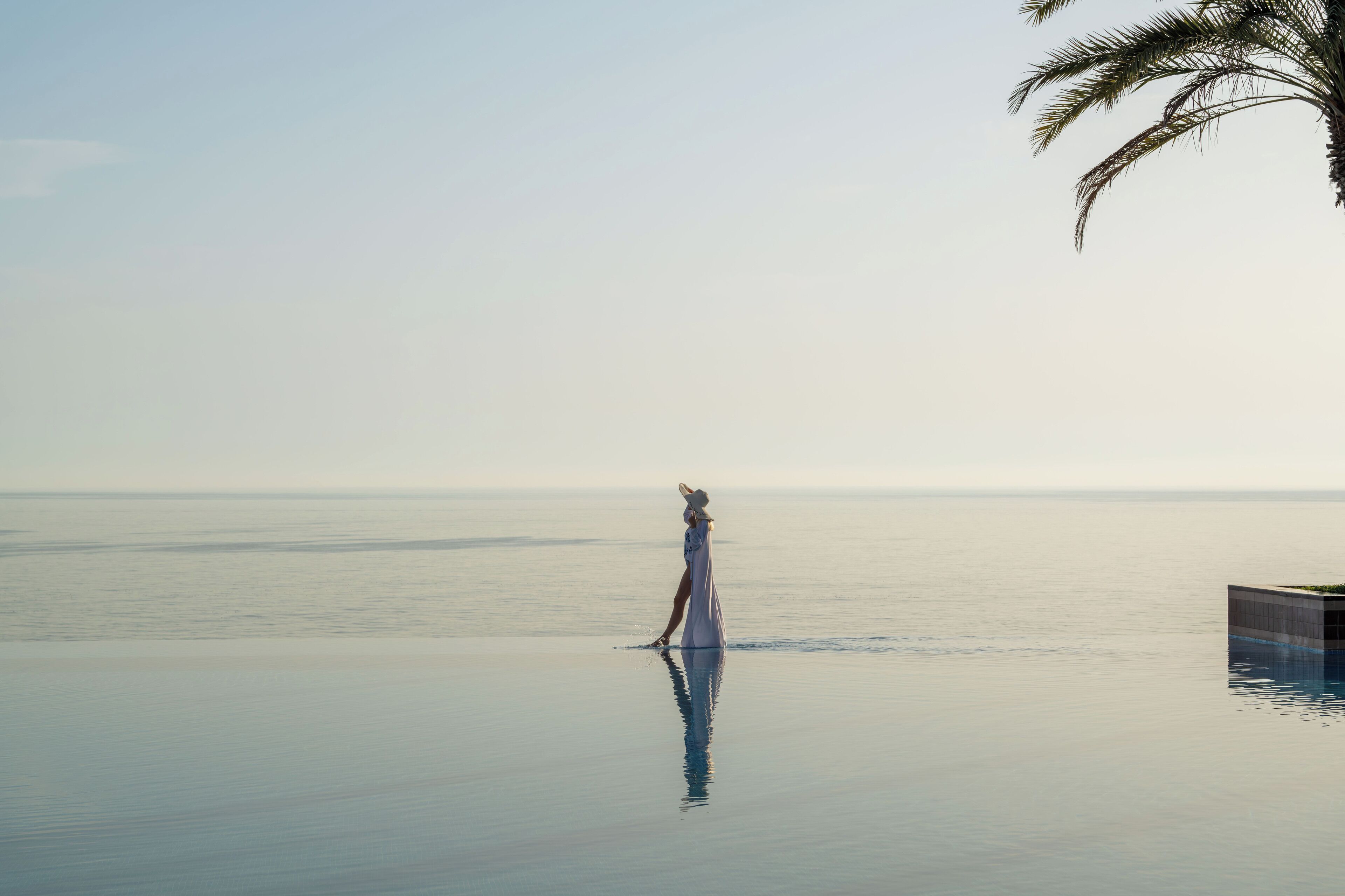 A person in a white dress standing on a glass-bottomed infinity pool with a view of the ocean and a palm tree in the background.
