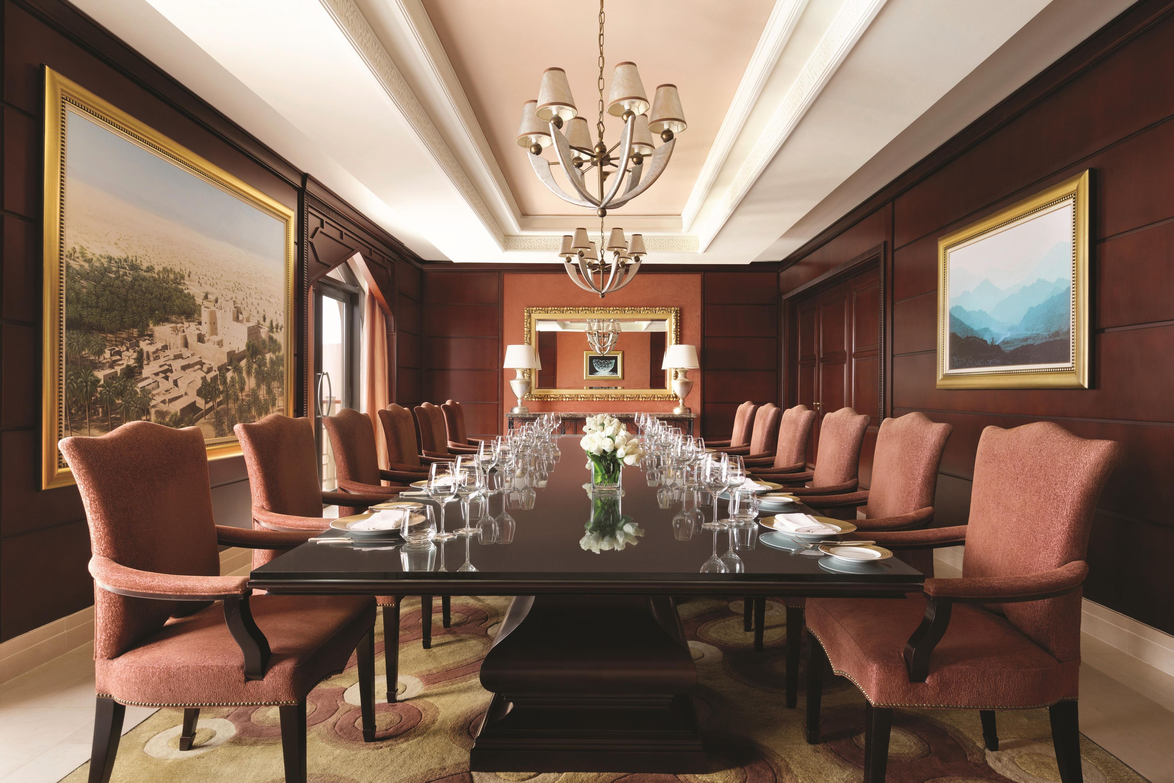 A formal dining room with a long table set for a meal, surrounded by upholstered chairs, a chandelier, and framed artwork on the walls.