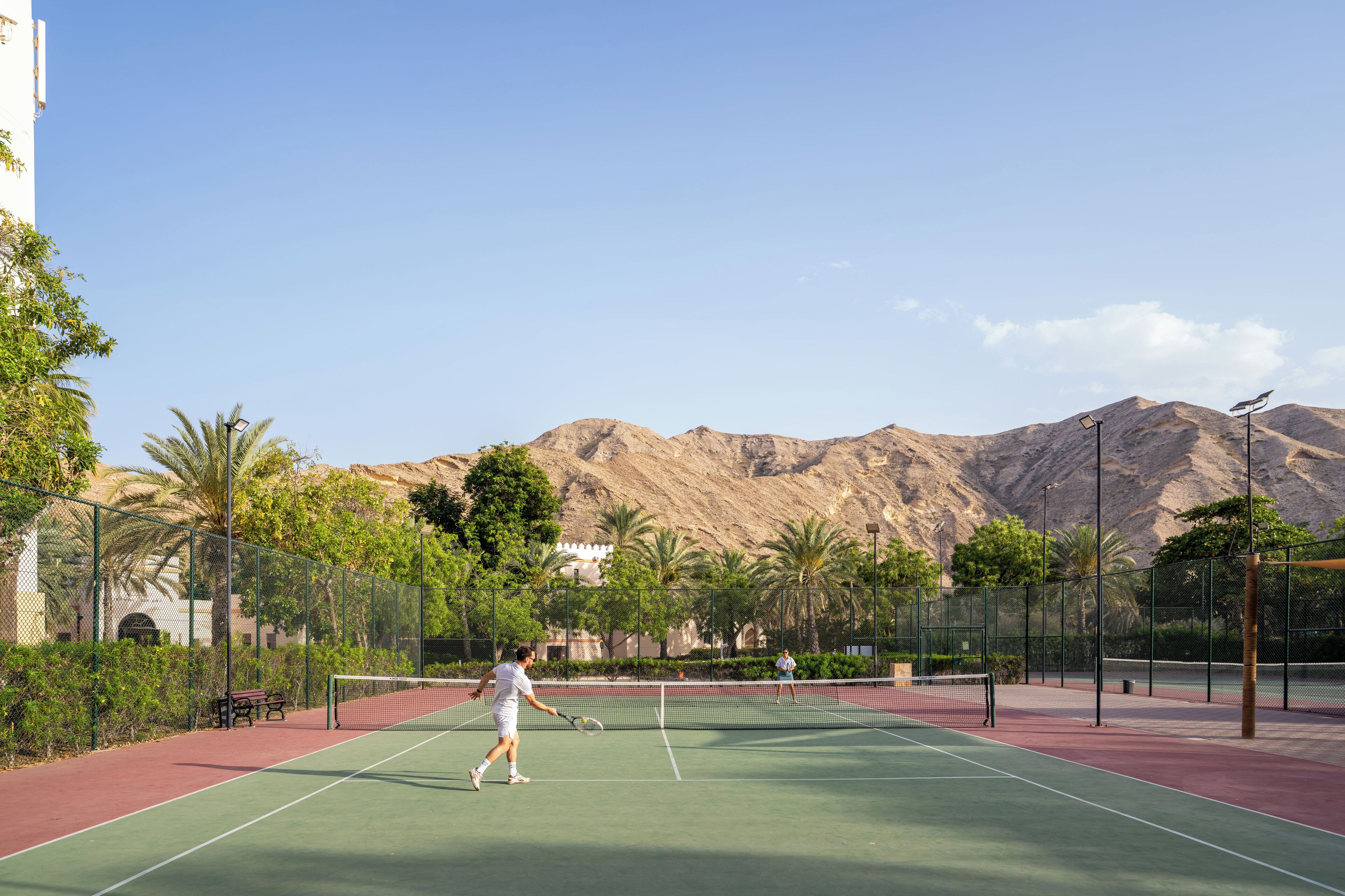 Una cancha de tenis con una red, rodeada por una valla, palmeras y montañas al fondo.