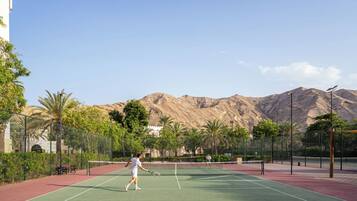 Una cancha de tenis con una red, rodeada por una valla, palmeras y montañas al fondo.