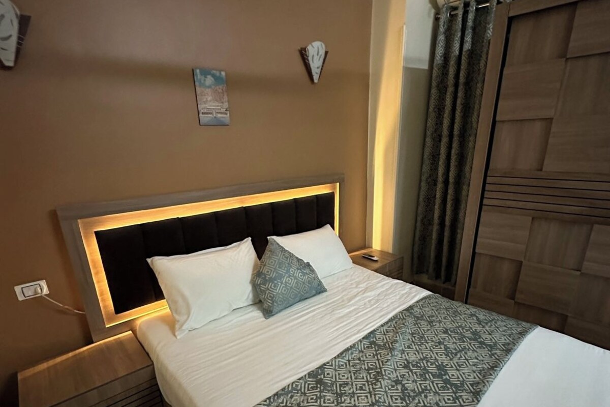 A neatly made bed with a patterned bedspread, a headboard with built-in lighting, a bedside table, and a wall-mounted clock.