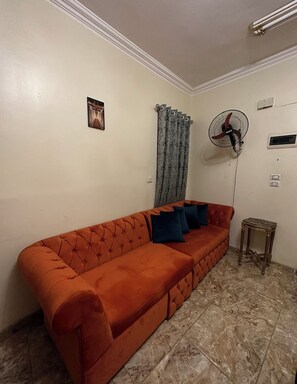 A living room with a red sofa, a patterned curtain, a standing fan, and a small table. - Luxe Nest (Luxor)