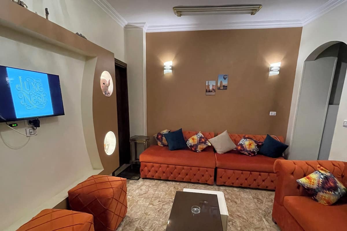A living room with a large orange sofa, matching ottomans, a flat-screen TV, a chandelier, and a fireplace.