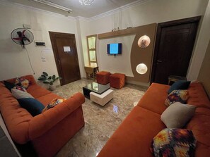 Una sala de estar con dos sofás naranja, una mesa de centro, un televisor y un ventilador de pared.