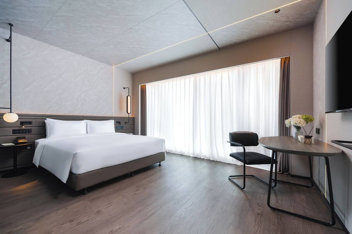 A modern hotel room with a large bed, bedside table, lamp, and a small table with flowers.
