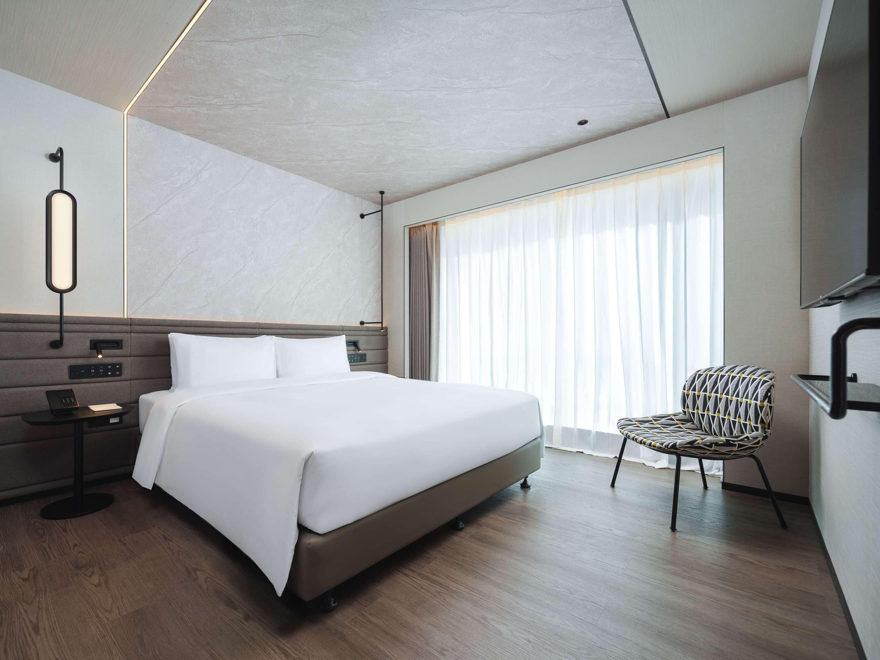 Una habitación de hotel moderna con una cama grande, una mesita de noche con un teléfono, una silla y un ventanal con cortinas.
