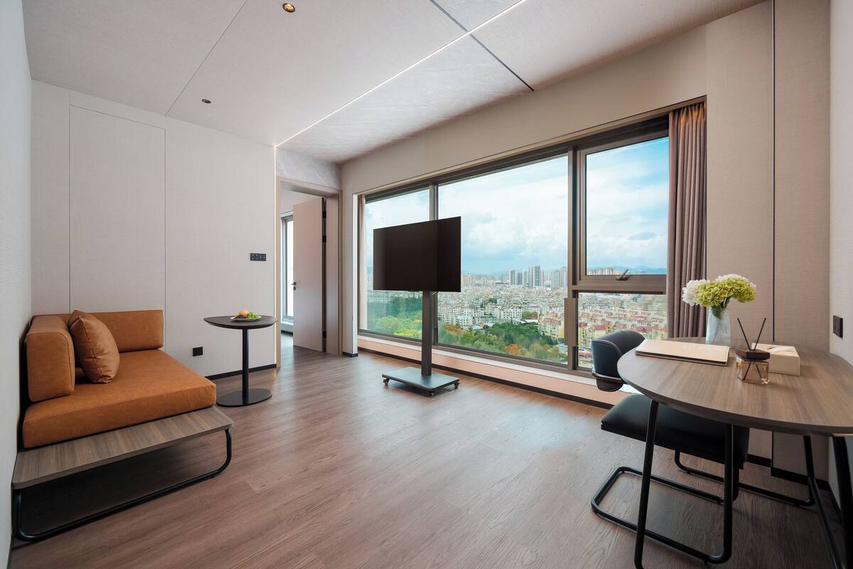 A modern hotel room with a large window, a sofa, a dining table, and a flat-screen TV.
