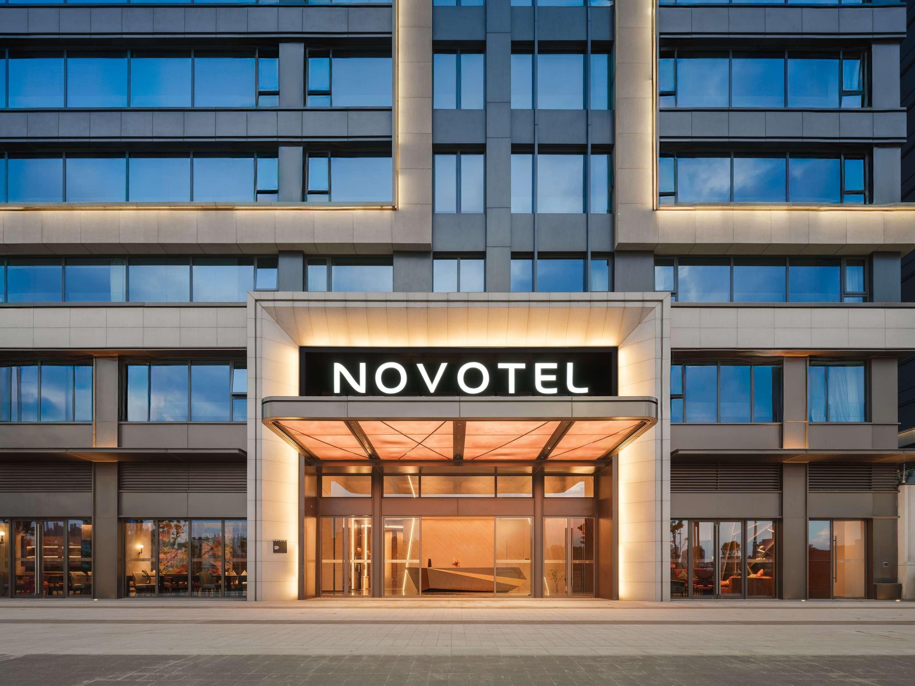 A modern hotel entrance with a glass facade, a prominent 'NOVOTEL' sign, and a well-lit entrance area.