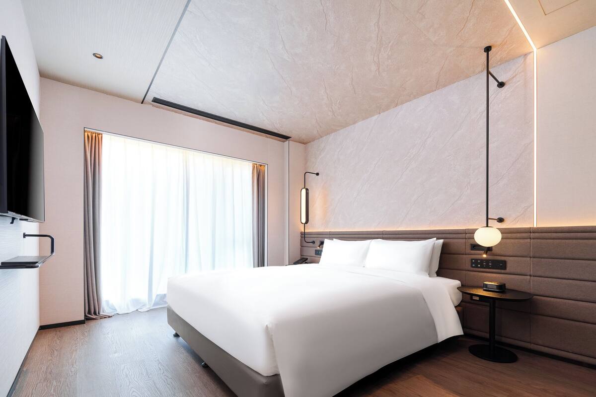 A modern hotel room with a large bed, a flat-screen TV, a bedside table, a lamp, and a large window with curtains.