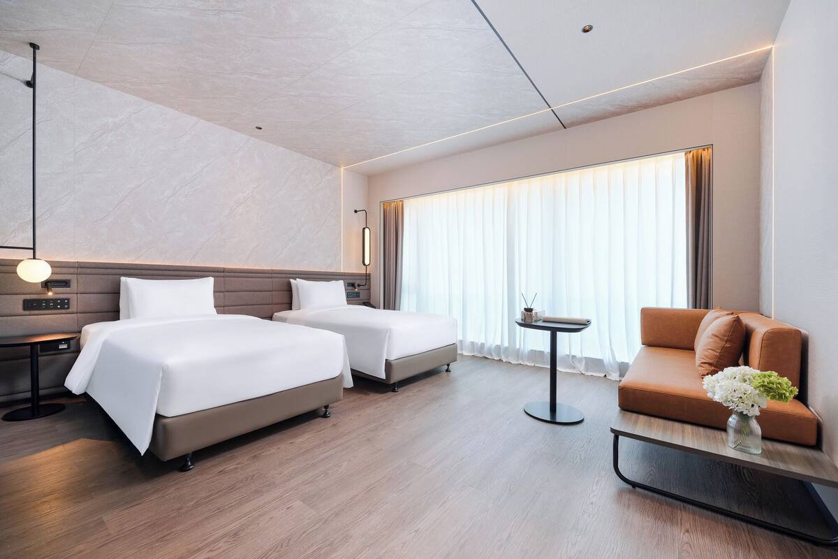 A modern hotel room with two beds, a sofa, a small table with a vase, and a large window with curtains.