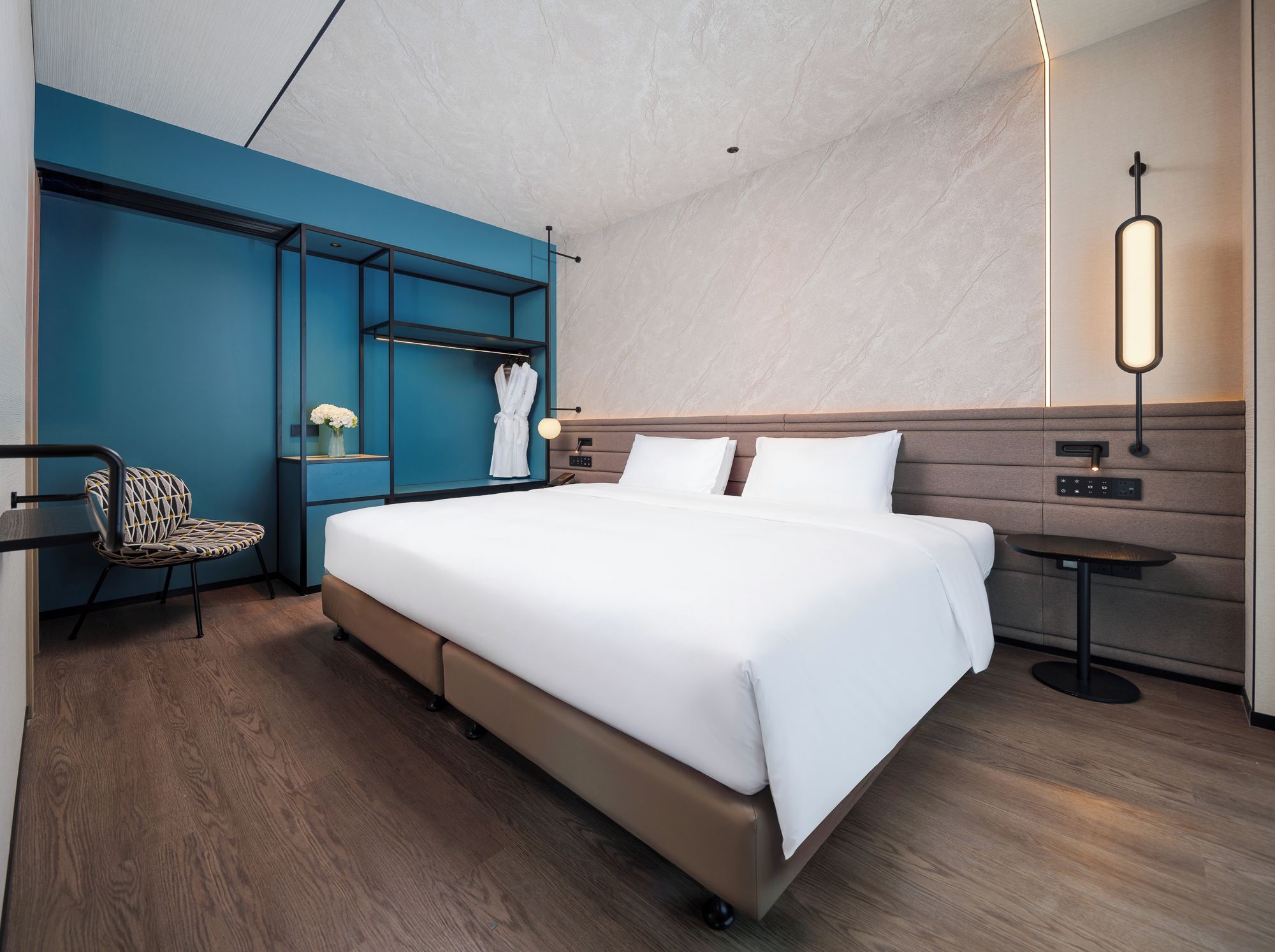 A modern hotel room with a large bed, a seating area with a chair, a robe rack, and a bedside table with a lamp.