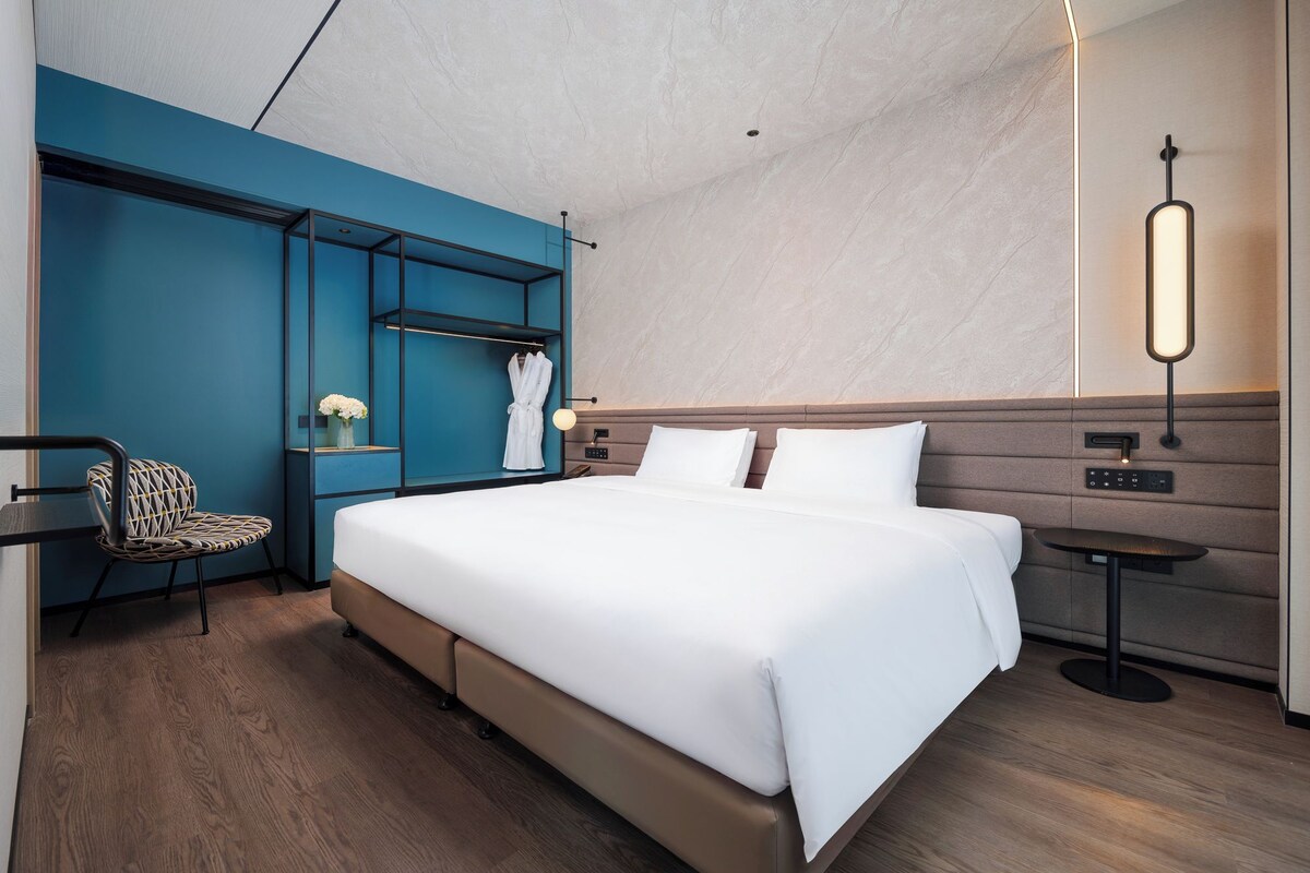 Une chambre d’hôtel moderne dotée d’un grand lit, d’un coin salon avec un fauteuil, d’un porte-peignoir et d’une table de chevet avec une lampe.