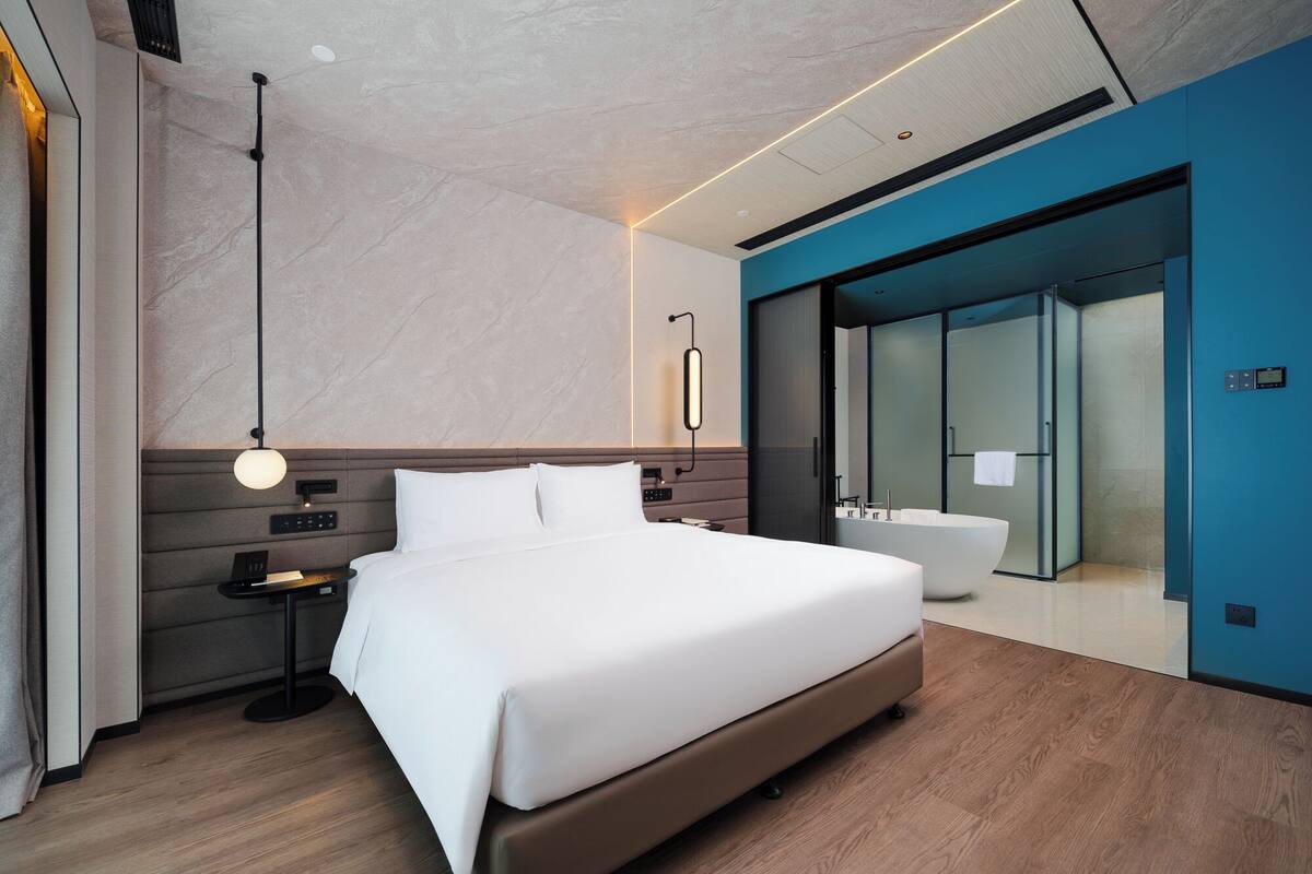 A modern hotel room with a large bed, bedside tables, a built-in bathtub, and a glass-enclosed shower area.