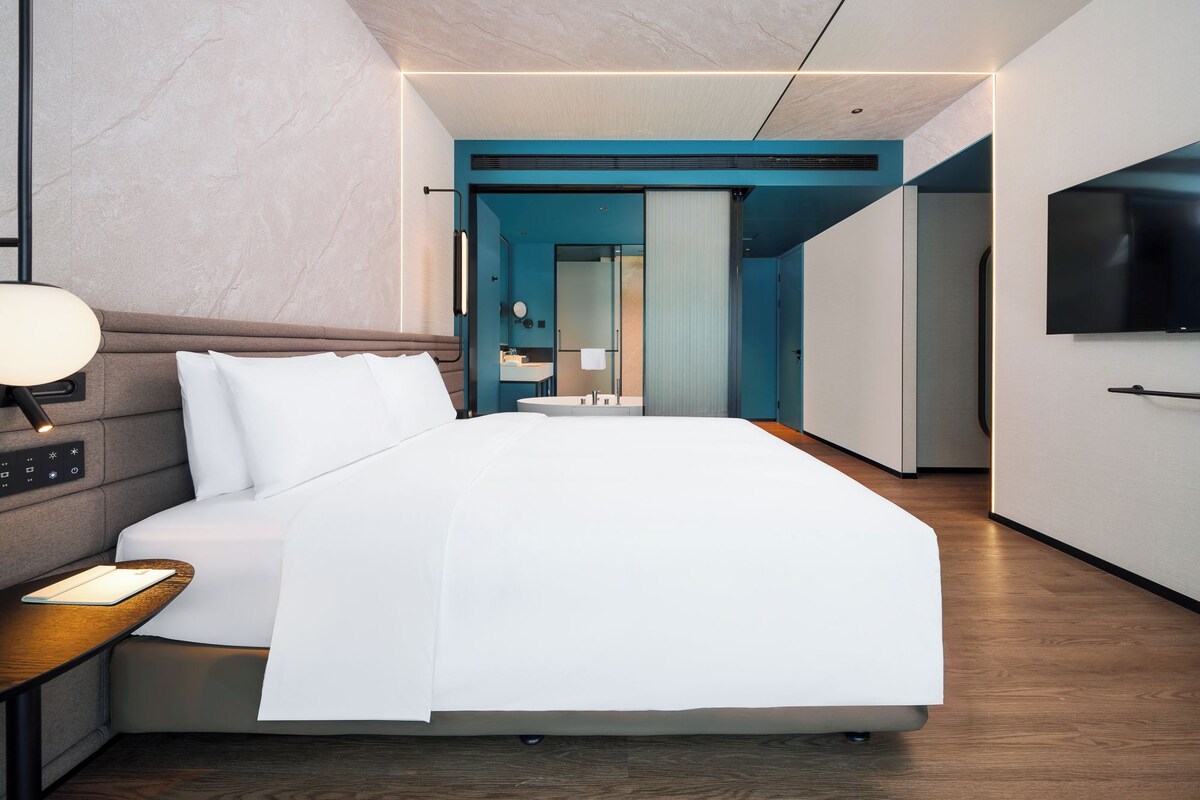 A modern hotel room with a large bed, bedside controls, a desk with a lamp, a TV, and a bathroom visible through an open door.