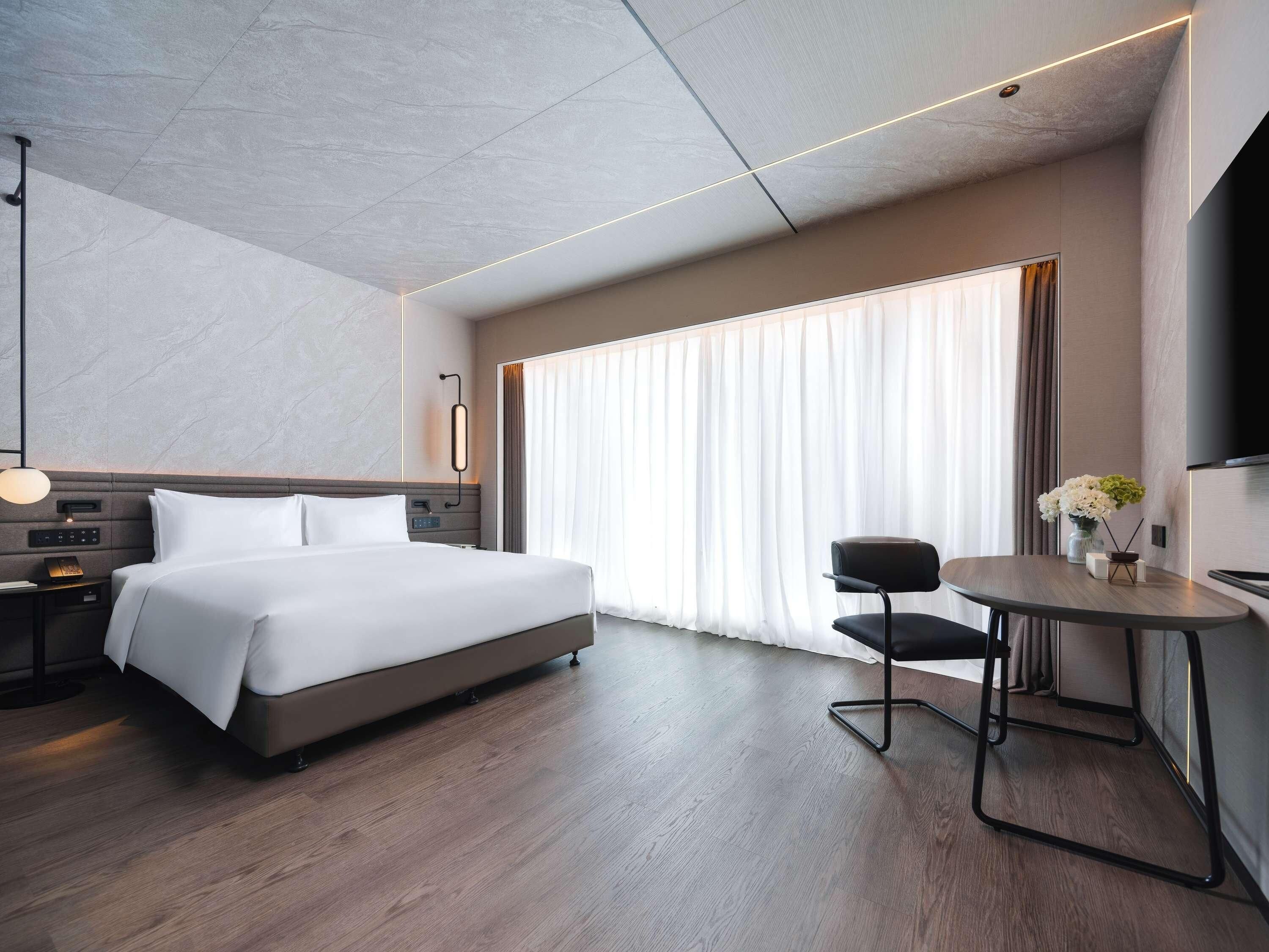 A modern hotel room with a large bed, a bedside table with a lamp, a small table with a chair, and a TV mounted on the wall.