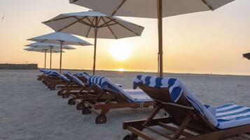 A row of sun loungers with striped towels and white umbrellas on a sandy beach at sunset.
