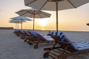 A row of sun loungers with striped towels and white umbrellas on a sandy beach at sunset. - Tulip Inn Majan Hotel (Salalah)