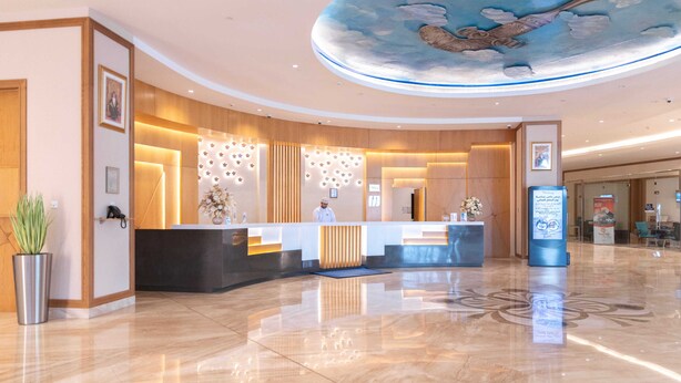A hotel reception area with a modern design, featuring a curved reception desk, a receptionist, and a ceiling mural. - Tulip Inn Majan Hotel (Salalah)