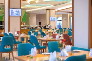 A hotel dining area with multiple tables and chairs, a buffet counter, and two televisions mounted on the walls. - Tulip Inn Majan Hotel (Salalah)