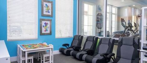 Ein Zimmer mit blauen Wänden, weißen Jalousien, einem Tisch mit bunter Tischdecke und Trainingsgeräten.