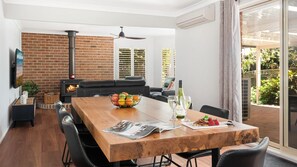 A dining area with a wooden table, black chairs, a bowl of fruit, wine glasses, and a magazine. There is a brick wall, a fireplace, and a view of greenery outside. - La Playa – your laid-back coastal getaway on the northern shores of Jervis Bay (Myola)