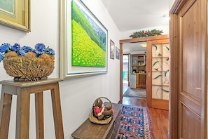 Um corredor com piso de madeira, tapete estampado, cesto de vime com um bicho de pelúcia e um quadro grande de um campo amarelo.