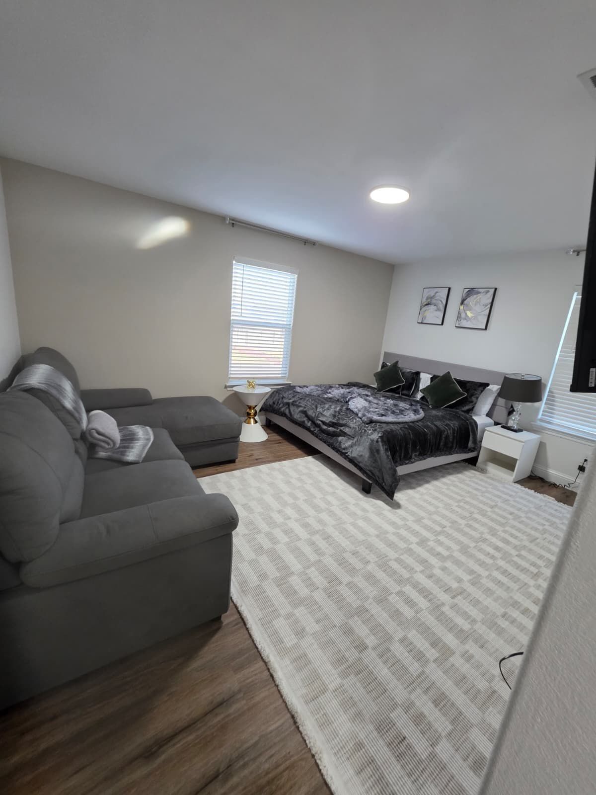 A modern bedroom with a bed, a grey sofa, a bedside table, and a window with blinds.