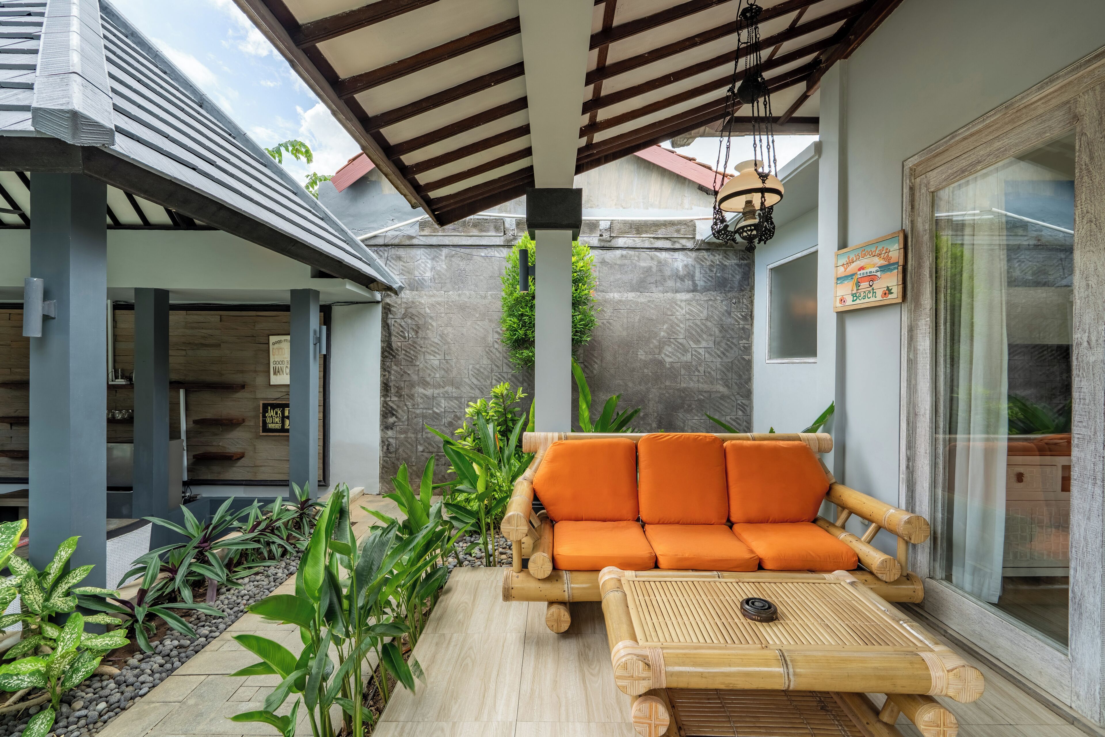 A covered outdoor seating area with a bamboo sofa and table, surrounded by plants and a stone wall.