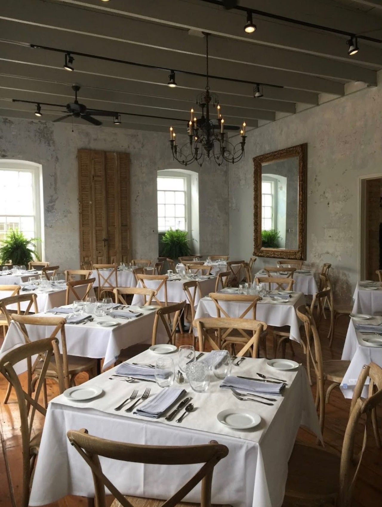 A dining area with tables set for service, wooden chairs, a chandelier, and a large mirror on the wall.