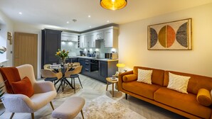 A modern living room with a sofa, armchairs, and a coffee table. There is a kitchen in the background with cabinets and appliances. - Micklegate Hideaway  -  Sleeps 4 Guests  in 2 Bedrooms (York)