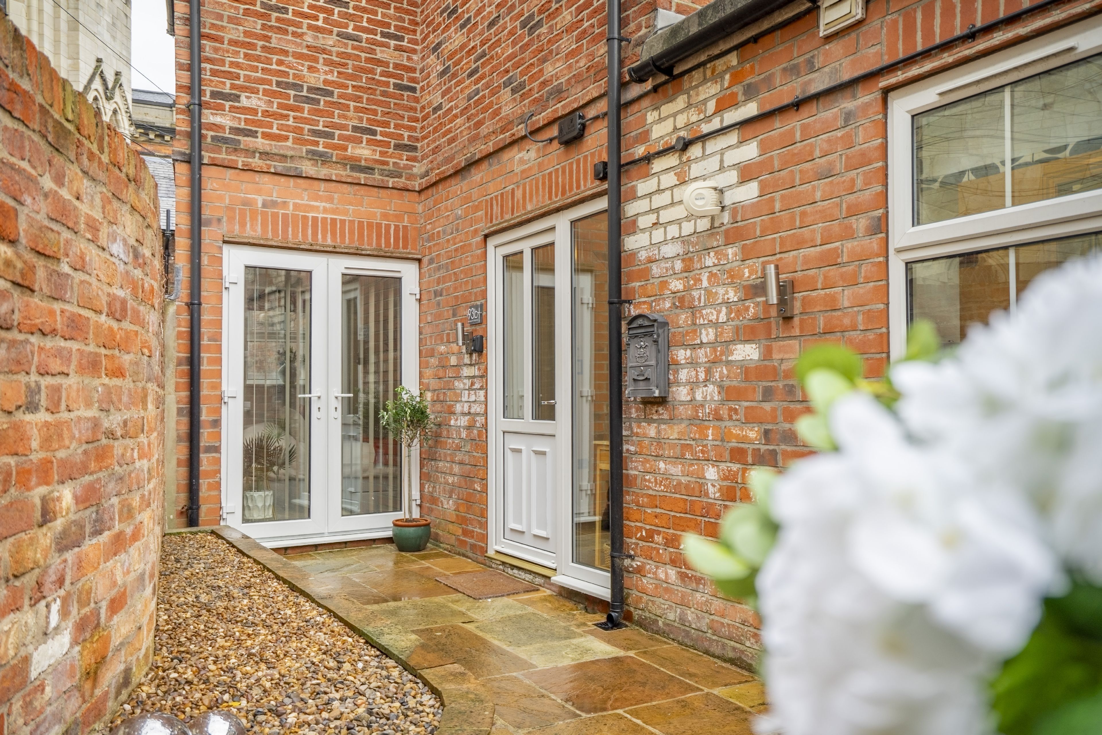 A brick building with a tiled patio, white doors, and a small garden area.