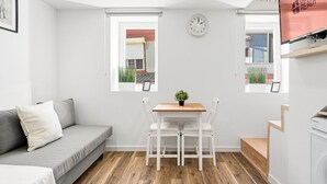 Una habitación moderna y compacta con un sofá gris, una pequeña mesa de madera con dos sillas, un reloj en la pared y una ventana con vistas a un edificio.