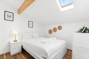 A minimalist bedroom with a bed, bedside table, and a nightstand with a lamp. There are three framed pictures on the wall, a ceiling with wooden beams, and a window with a view.