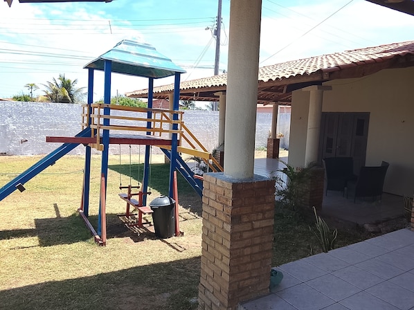 Un parque infantil con una estructura de escalada azul y amarilla, un muro de ladrillo y una zona de estar cubierta con sillas.