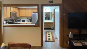 Eine kleine Küche mit Holdekor-Schränken, einer Mikrowelle und einem Kühlschrank. Ein Essbereich mit Tisch und Stühlen ist zu sehen, und an der Wand ist ein Fernseher angebracht.