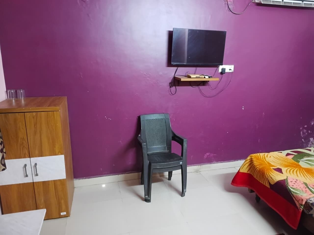 Een kamer met paarse wanden, een houten kast, een zwarte plastic stoel, een bed met een bloemenprint en een aan de muur bevestigde televisie.
