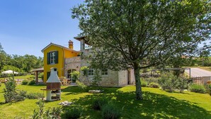 Une maison jaune aux murs en pierre, dotée d’un barbecue en briques et d’un jardin bien entretenu avec un grand arbre.